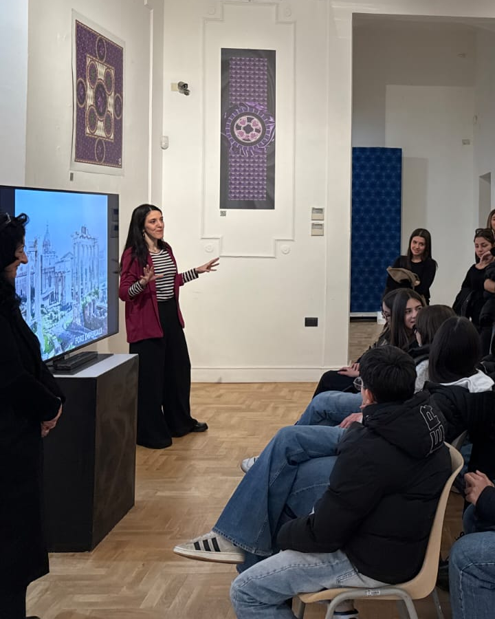 Francesca Librandi parla al pubblico in una sala espositiva, accanto a uno schermo con un'immagine dei Fori Imperiali